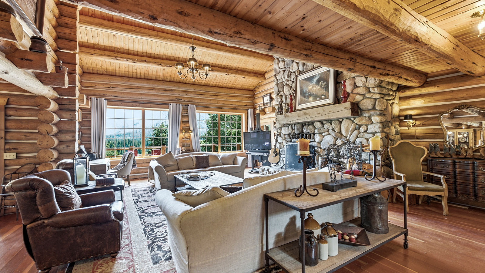 Log cabin living room with a leather sofa, reamed coffee table, vaulted wood ceiling, and large windows looking out to green trees and mountains.