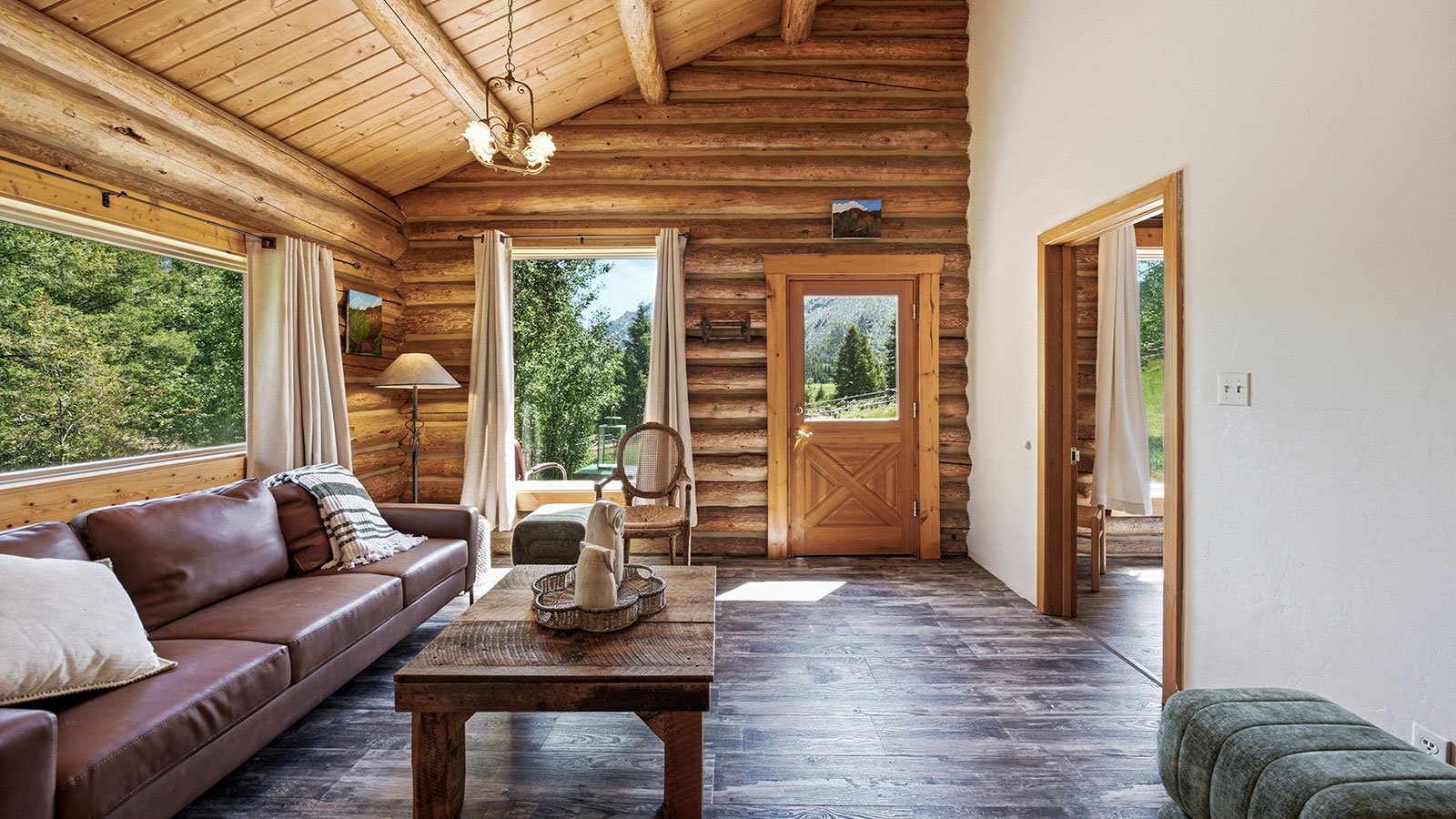 Log cabin great room with a tall river-rock fireplace, vaulted wood ceiling, sofas, leather chairs, and large windows over the forest.