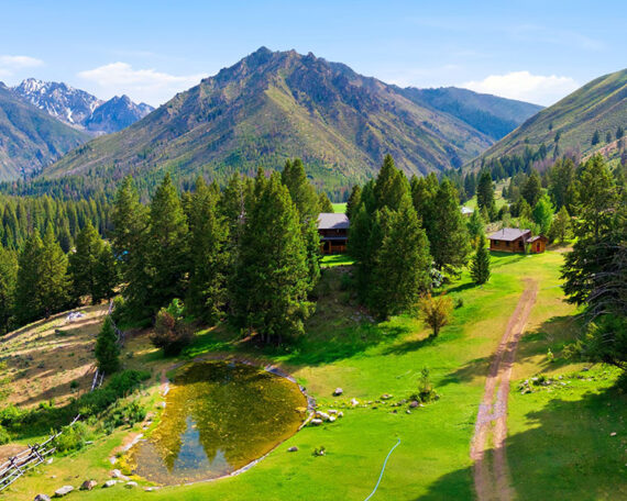 Aerial view of Loon Creek Retreat's lodge and cabin among conifers, with a pond and pasture below the Salmon River Mountains.